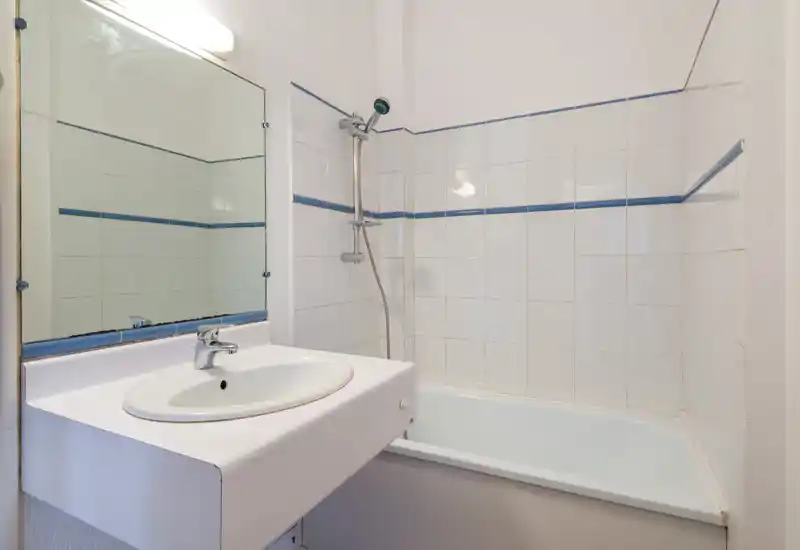 Compact bathroom with white tiled walls, bathtub and wall-mounted shower head; simple sink with mirror above, bright neutral lighting.