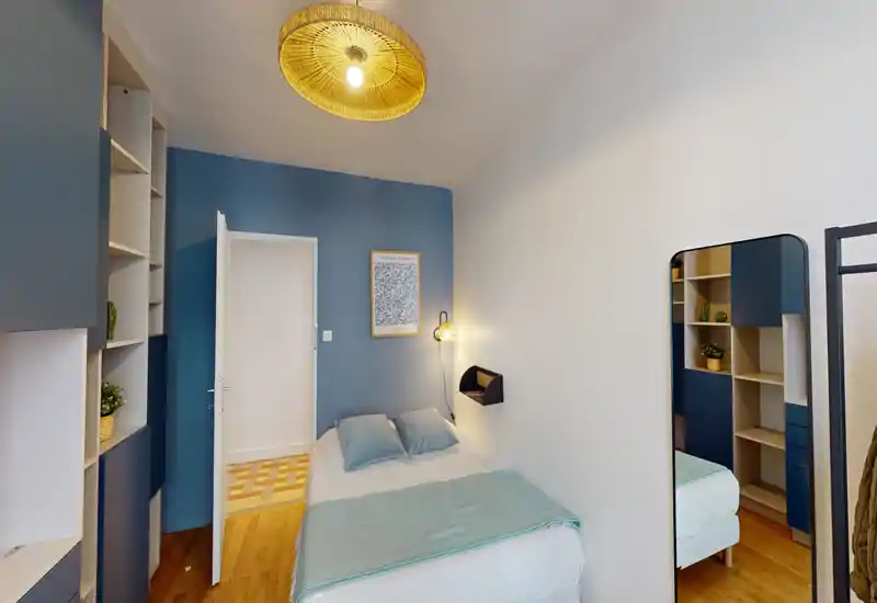 Bright, well-styled small bedroom with a double bed, soft blue accents and built-in shelving. Warm pendant light and wall lamp create a cozy atmosphere, and a full-length mirror plus wooden floor add depth.