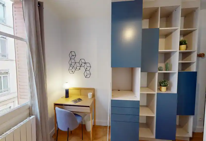 Bright, modern bedroom corner with a study desk, lamp and built-in shelving; large window provides natural light and the space is styled with small plants and decorative details.