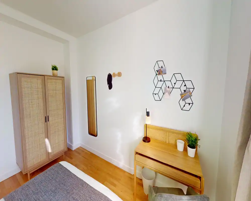 Coin chambre lumineux et minimaliste avec lit partiellement visible, armoire en rotin, petit bureau en bois et étagère décorative — matériaux naturels et lignes épurées pour une ambiance calme et accueillante.