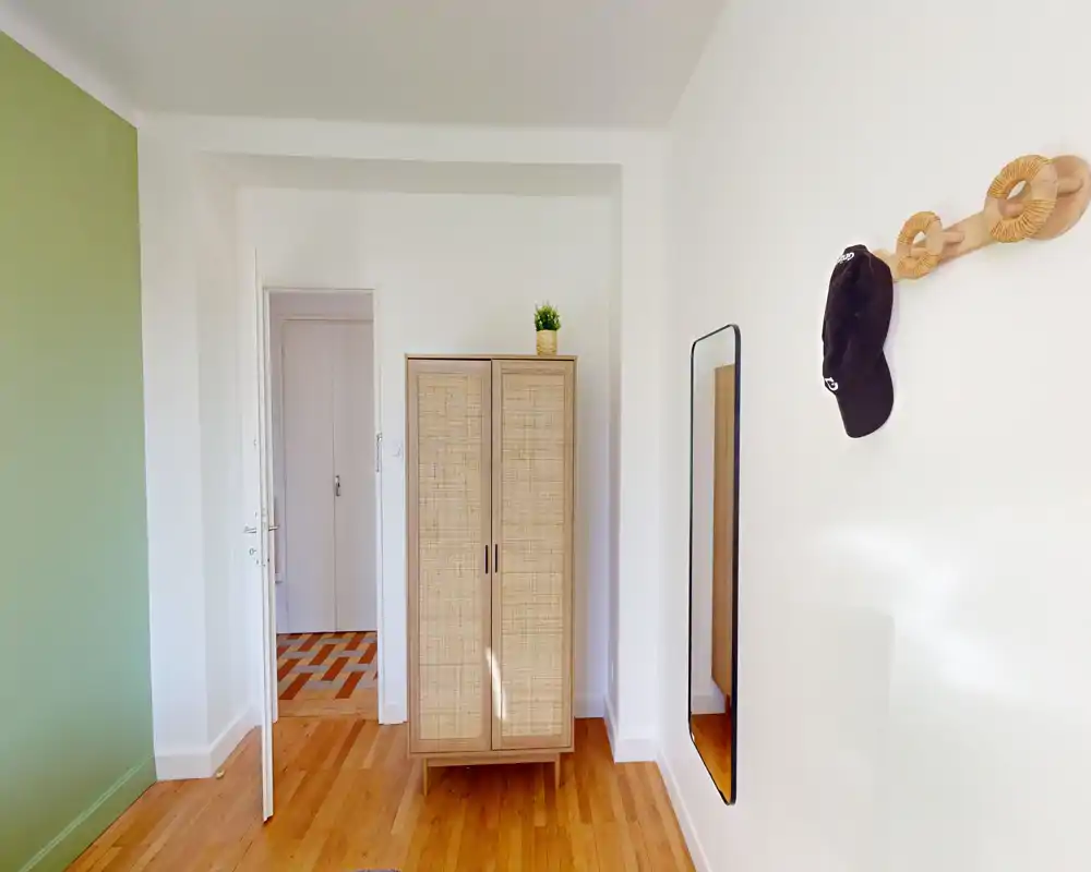 Pièce lumineuse et minimaliste avec une armoire en rotin, un miroir pleine longueur et des crochets en bois — espace neutre et bien éclairé, adapté à une chambre simple ou un petit studio.