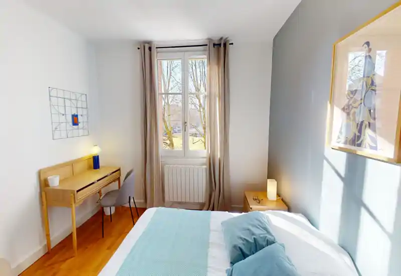 Sunny, well-styled bedroom with a comfortable double bed, soft blue textiles and a light wooden desk by the window — ideal for relaxing or working. Natural light fills the space through a large window with neutral curtains.