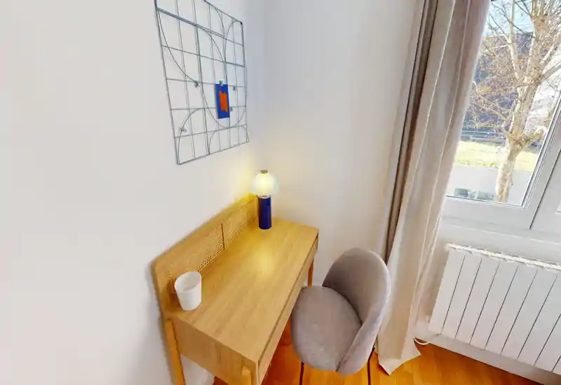 Small bright bedroom corner with a wooden desk, upholstered chair and bedside-style lamp next to a large window letting in natural light.