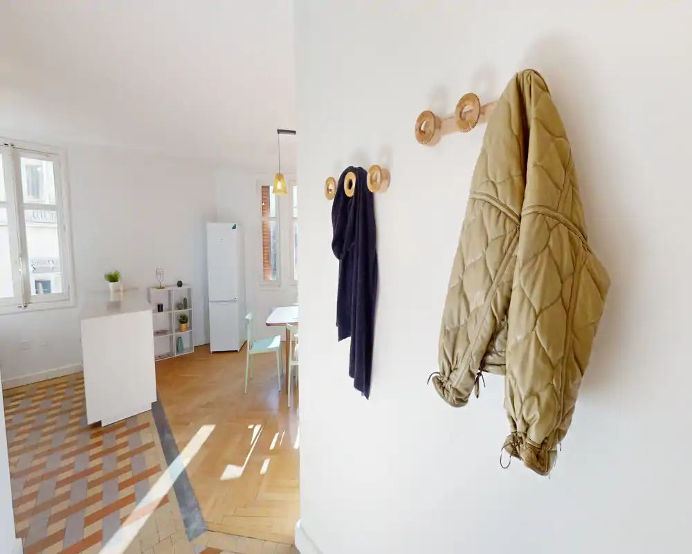 Bright, minimal entrance/foyer with wall hooks holding a jacket and scarf, leading into an open-plan living/dining area with natural wood flooring and tiled entryway.