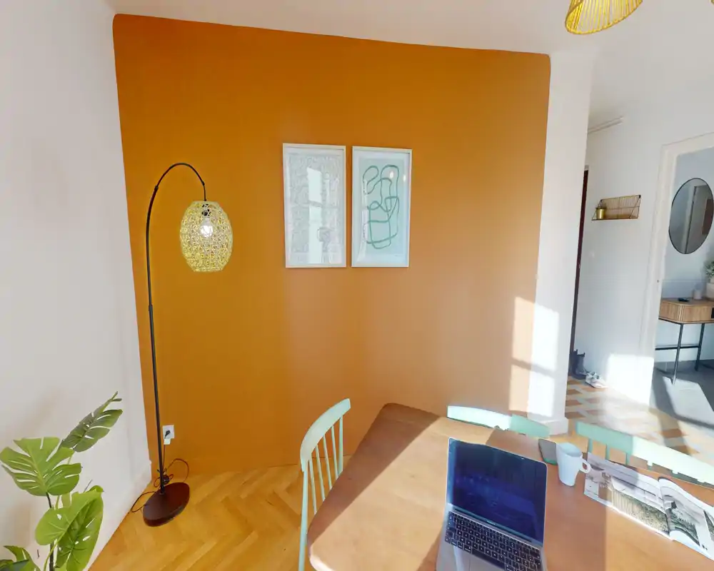 Bright, warm dining corner with an orange accent wall, wooden table, and mint-green chairs — ideal for relaxed meals and daytime work by the window.