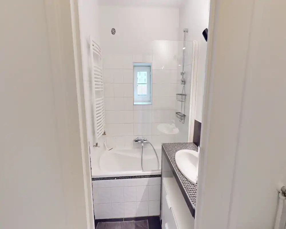 Bright, modern bathroom with a combined bathtub and shower, white tiled walls, a small window and a sink with countertop storage — neat and functional.