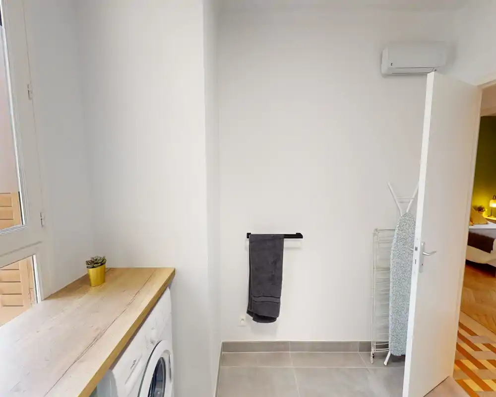 Small, functional laundry/utility area with a countertop, front-load washer, towel rail and ironing board. Natural light from the window keeps the space fresh and practical.