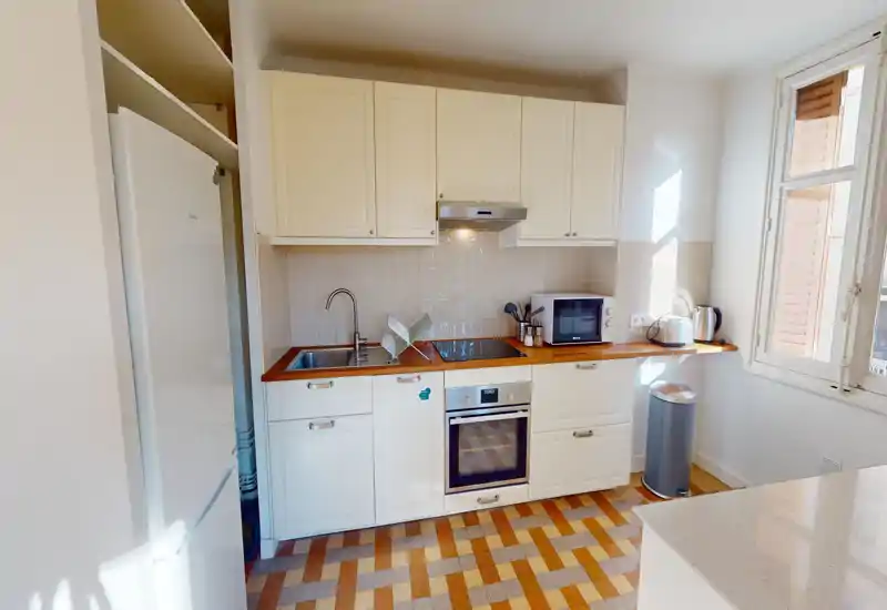 Cuisine compacte et lumineuse avec meubles blancs, plan de travail en bois, four encastré et plaque à induction. La lumière naturelle par la fenêtre rend l'espace accueillant et adapté aux locations courtes durées.