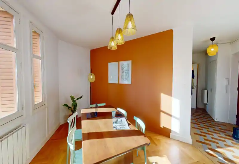 Coin repas lumineux et aménagé avec table en bois, chaises pastel et suspensions chaleureuses devant un mur orange — idéal pour des annonces valorisant un coin repas convivial.