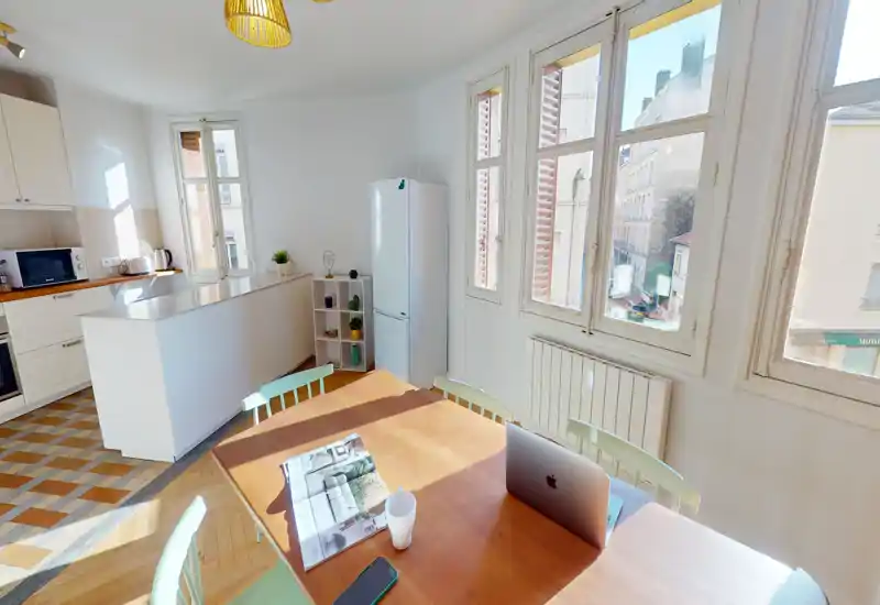 Cuisine ouverte lumineuse avec table en bois baignée de soleil, meubles blancs, un bar et de grandes fenêtres donnant sur la rue — idéale pour un repas convivial ou le télétravail.