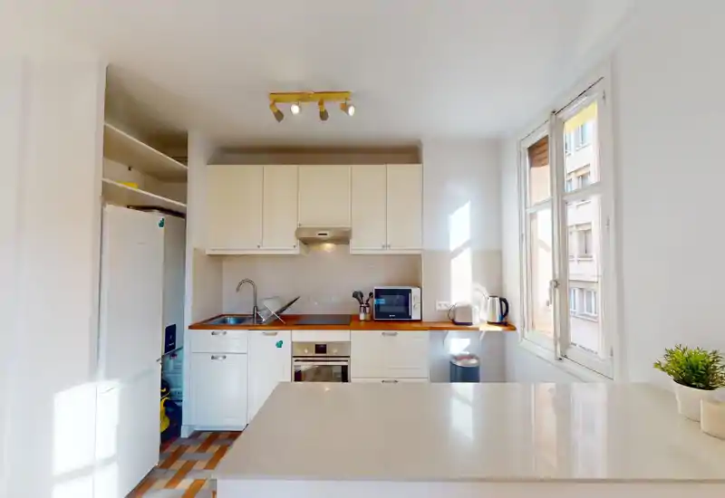Cuisine compacte et lumineuse avec placards blancs, plan de travail en bois et électroménagers modernes (four, micro-ondes, réfrigérateur); un bar donne sur une grande fenêtre très lumineuse.