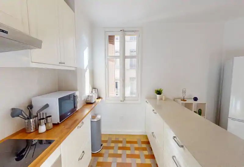 Cuisine moderne et lumineuse avec meubles blancs, plan de travail en bois, bar et sol carrelé décoré ; la lumière naturelle et la décoration soignée rendent l’espace chaleureux.