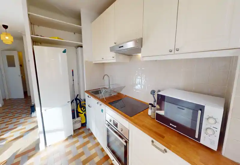 Cuisine compacte et lumineuse avec plan de travail en bois, four encastré, plaque à induction et micro-ondes. Espace propre et bien éclairé avec placards blancs et crédence carrelée.