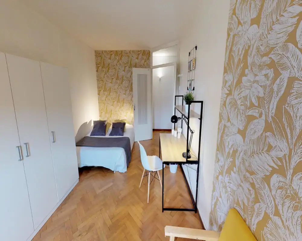 Dormitorio luminoso y bien decorado con cama doble, cojines y una zona de trabajo. Papel pintado cálido y suelo de parquet crean una sensación acogedora y moderna, perfecto para un anuncio urbano.