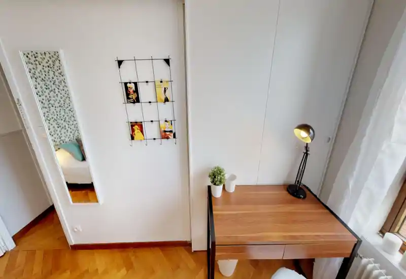 Bright, modern bedroom corner shown with a wooden desk and a lamp; a mirror at left reflects a patterned-wall bedroom with pillows and part of the bed.