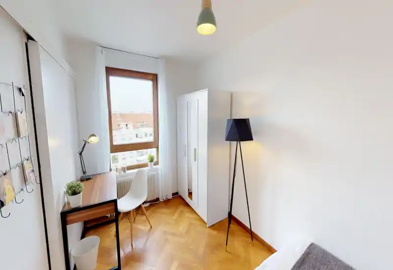 Bright, minimalist bedroom corner with a small desk by the window, white wardrobe and a floor lamp. The bed is partially visible in the foreground, and warm parquet flooring creates a cozy atmosphere.