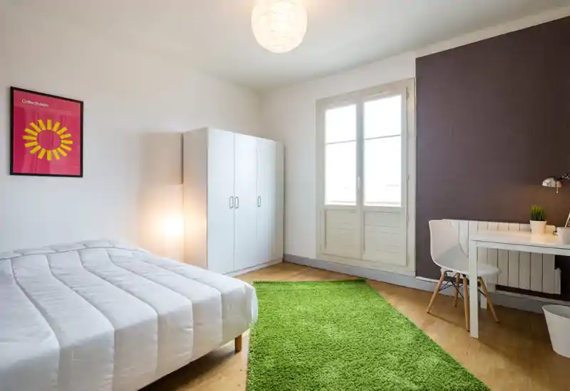 Bright, minimalist bedroom with a neatly made double bed, white wardrobe, desk area and a vivid green rug underfoot. Large window provides ample natural light and the room is staged simply for a fresh, airy feel.