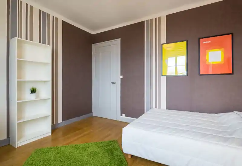 Bright, modern bedroom with a white bed, patterned accent wall and simple shelving; wooden floors and a green rug add warmth.