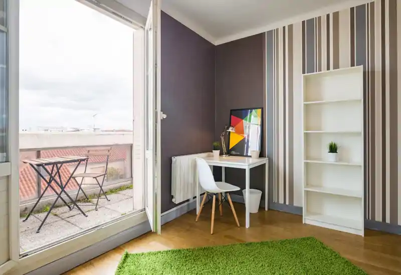 Bright, well-styled living corner with a small desk, chair and bookshelf opening onto a balcony. Natural light and minimalist decor make this space welcoming for work or relaxation.