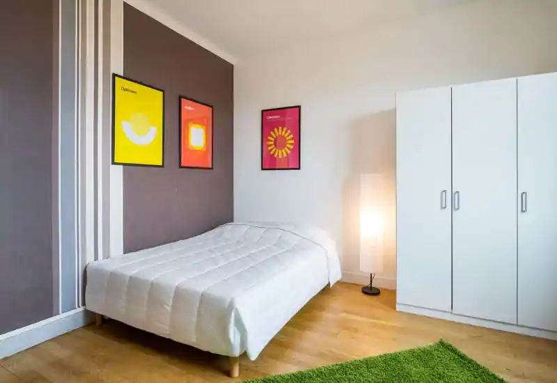 Bright, minimal bedroom with a neatly made double bed, modern framed artwork and a white wardrobe. The room is simply styled with warm lighting and wooden floors, ideal for a rental listing highlighting cleanliness and calm.