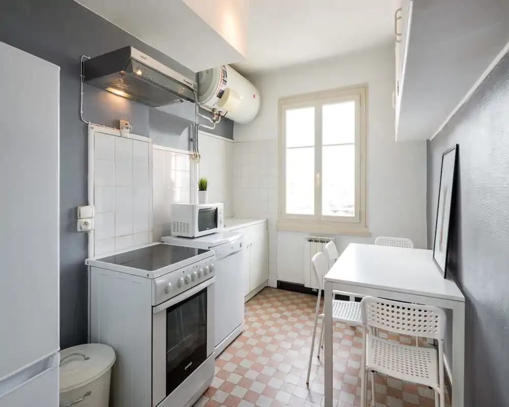 Bright, compact kitchen with a freestanding stove, microwave and a small dining table next to a large window; clean, minimalist styling and good natural light.