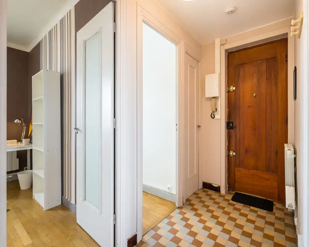 Bright apartment entrance with a wooden front door, patterned tiled floor and visible intercom; doorway opens to adjoining rooms with parquet flooring.