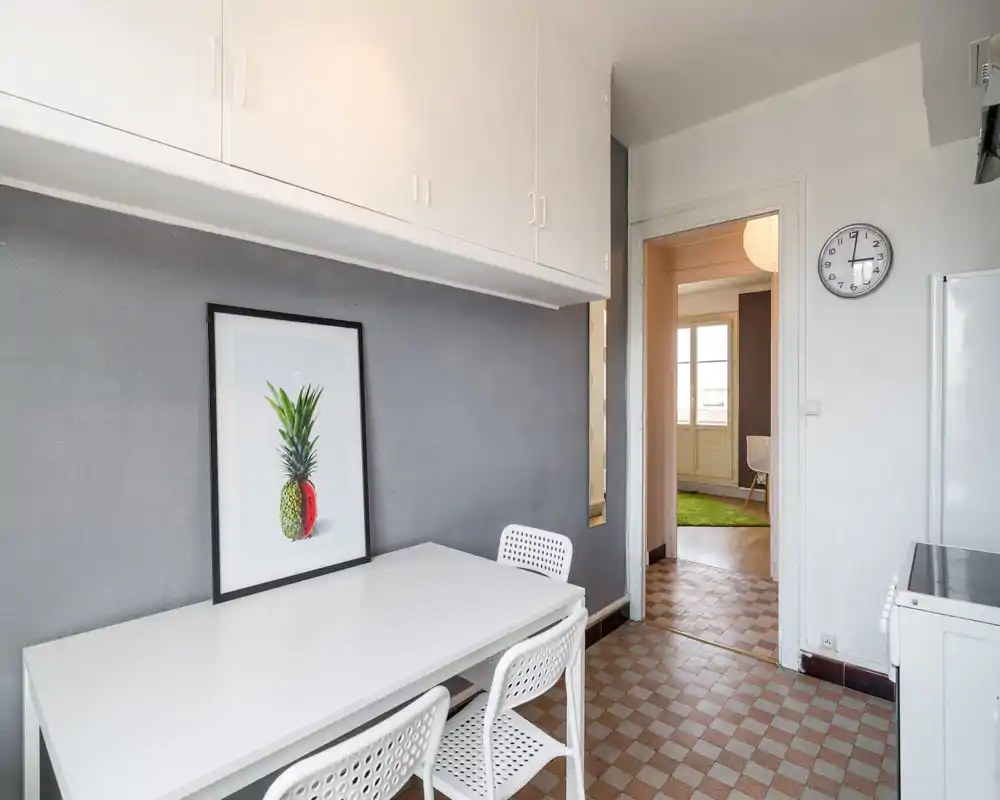 Bright, tidy kitchen nook with a white table and chairs, overhead cabinets and simple decorative artwork; clean, functional space with natural light from the adjacent room.