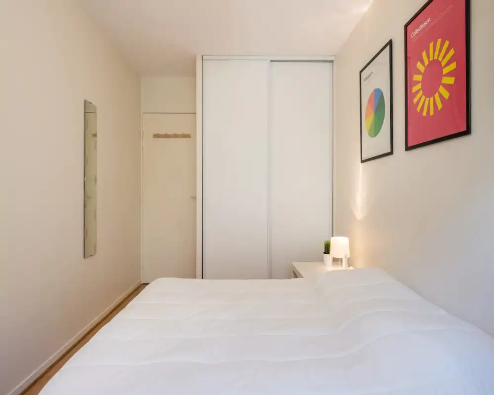 Bright, minimalist bedroom with a large white bed, sliding wardrobe doors and simple framed art; a bedside table with a lamp adds a cozy touch.