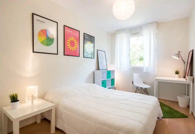 Bright, modern bedroom with a large white bed, minimalist desk and natural light from a window. Clean Scandinavian styling with framed prints, green accents and cozy lighting.