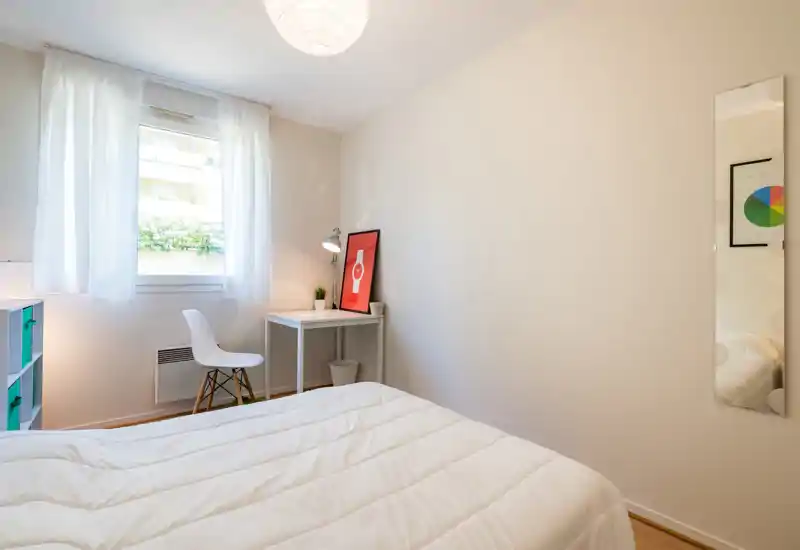 Bright, minimalist bedroom with a white quilted bed in the foreground, a small desk and chair by the window, and simple decorative accents for a calm, airy feel.