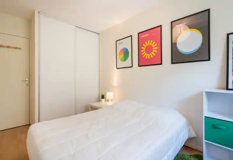 Bright, minimal bedroom with a large white bed, bedside table with lamp and plant, framed artwork on the wall and built-in sliding wardrobe — modern, clean and welcoming.