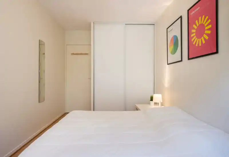 Bright, minimalist bedroom with a large white bed, sliding wardrobe doors and simple framed art; a bedside table with a lamp adds a cozy touch.