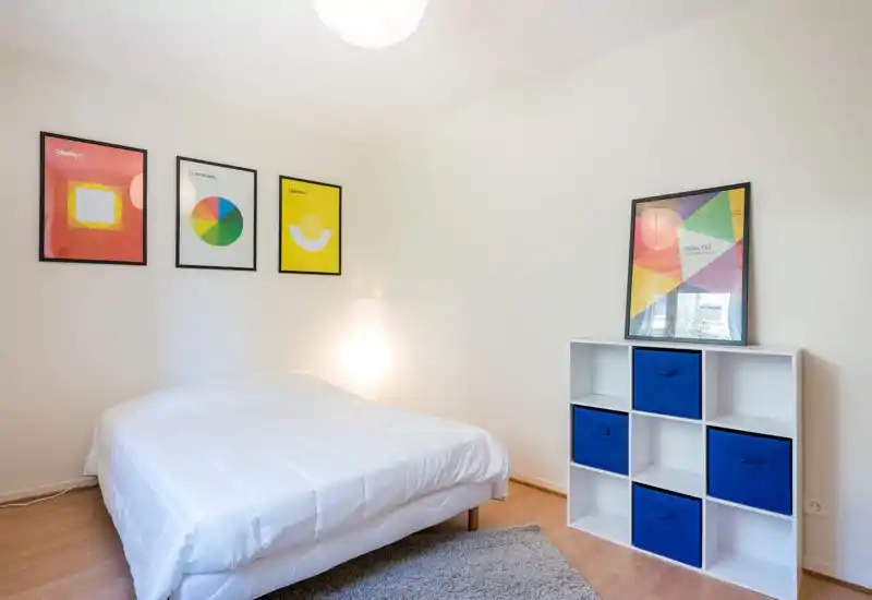 Bright, minimal bedroom with a neatly made double bed, colorful framed prints on the wall and a white cube storage unit with blue bins — calm, modern and suitable for rental listings.