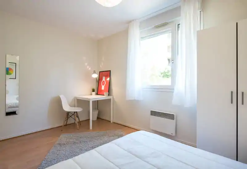 Bright, minimalist bedroom with a white bed in the foreground, a small desk and lamp near a large window that fills the room with natural light.