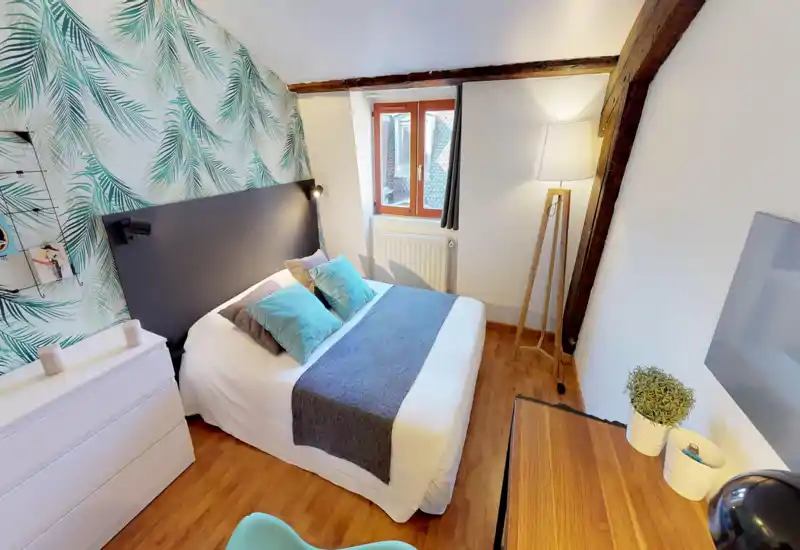 Cozy, well-styled bedroom with a double bed, patterned feature wall and wooden floor. Natural light from the window plus a floor lamp create a bright, welcoming atmosphere—ideal for short stays.