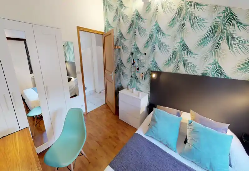 Bright, stylish bedroom with a double bed, decorative cushions and leafy wallpaper. The room features a wardrobe with mirrored doors, a chest of drawers and a teal accent chair.