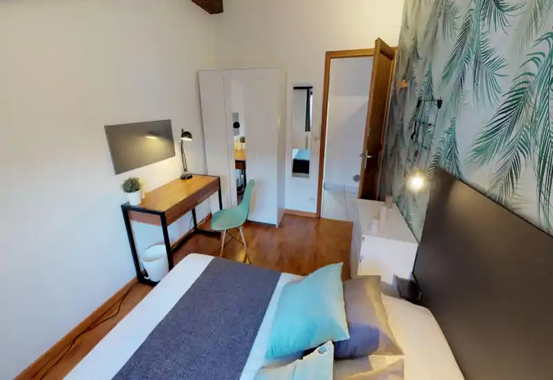 Bright, stylish bedroom with a comfortable double bed in the foreground, decorative pillows and a patterned feature wall. Includes a small workspace with desk and chair and a wardrobe — ideal for a cozy short-term stay.