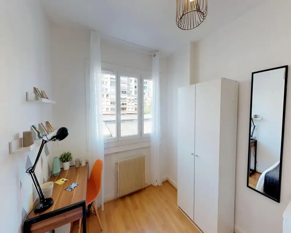 Dormitorio luminoso y ordenado con zona de estudio: escritorio de madera con lámpara y silla naranja junto a una gran ventana, armario y espejo de cuerpo entero que refleja parte de la cama.