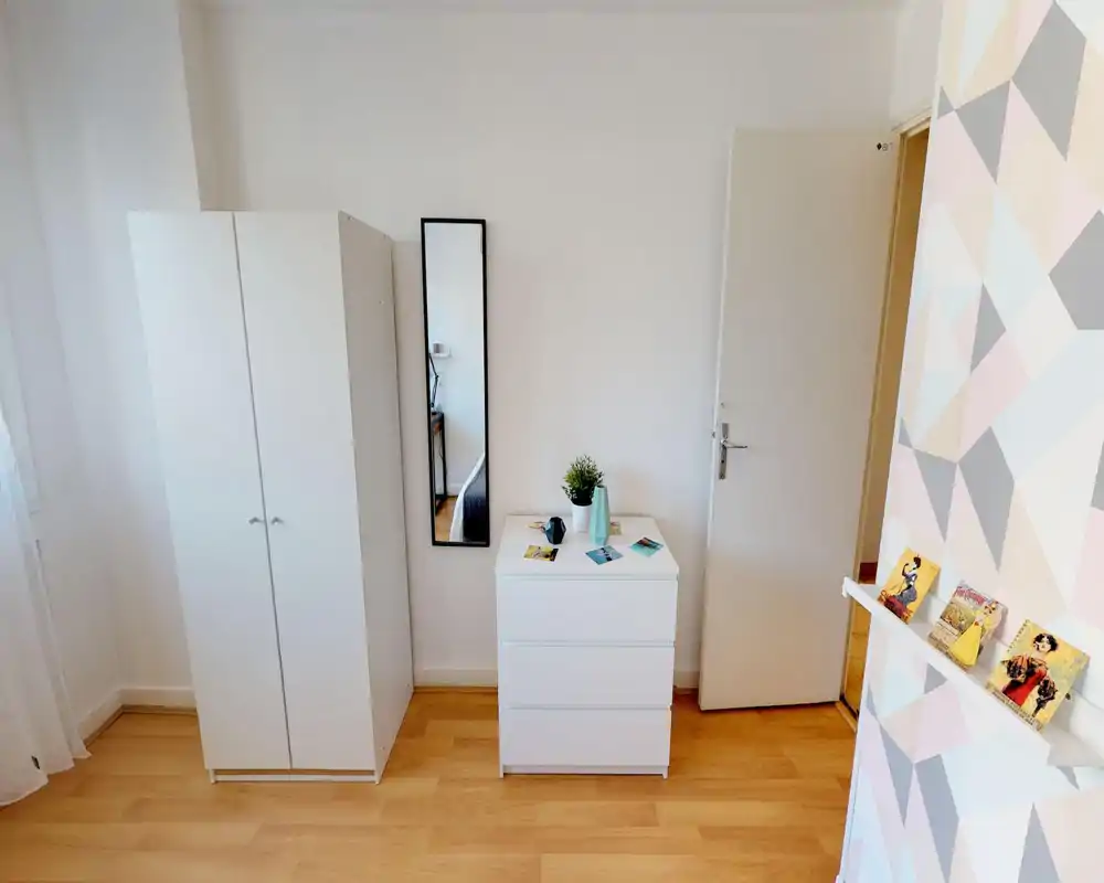 Rincón de dormitorio luminoso y minimalista con armario blanco, cómoda de tres cajones y espejo vertical que refleja parte de la cama; decoración sencilla y suelo de madera.