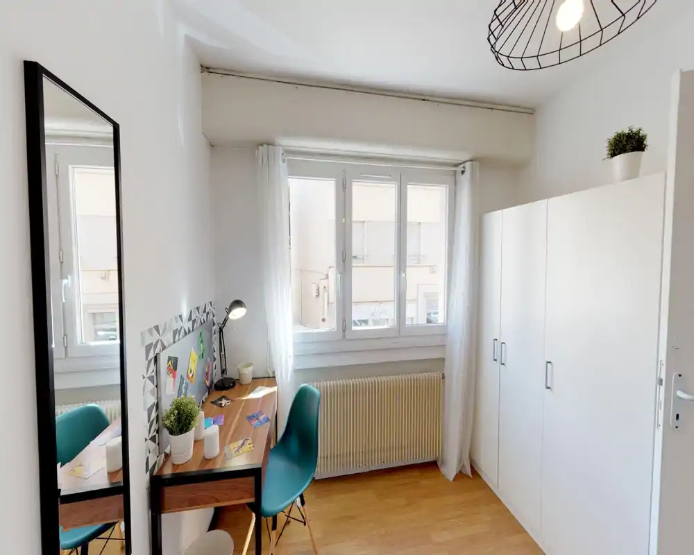 Camera da letto luminosa e compatta con una scrivania vicino alla finestra, ampia armadiatura e stile moderno minimal. Le tende leggere e il pavimento in legno creano un’atmosfera fresca e accogliente.