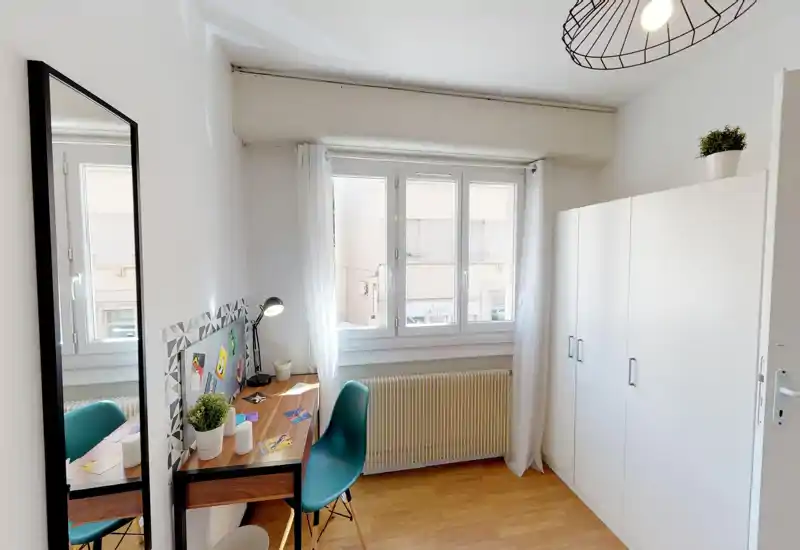 Bright, compact bedroom with a studied workspace by the window, large wardrobe and modern minimal styling. Sheer curtains and wooden flooring create a fresh, welcoming atmosphere.