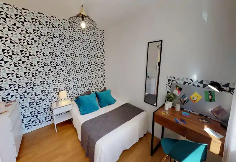 Bright, stylish bedroom with a patterned accent wall, a neatly made double bed with teal cushions, bedside table and a practical wooden desk by the window.
