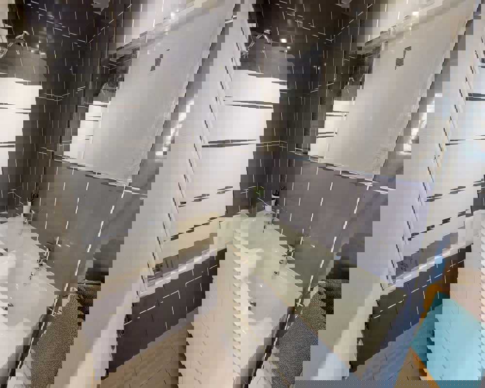 Bagno moderno e pulito con vasca/doccia, mobile con doppi lavelli e grande specchio; piastrelle grigie e disposizione compatta e luminosa.