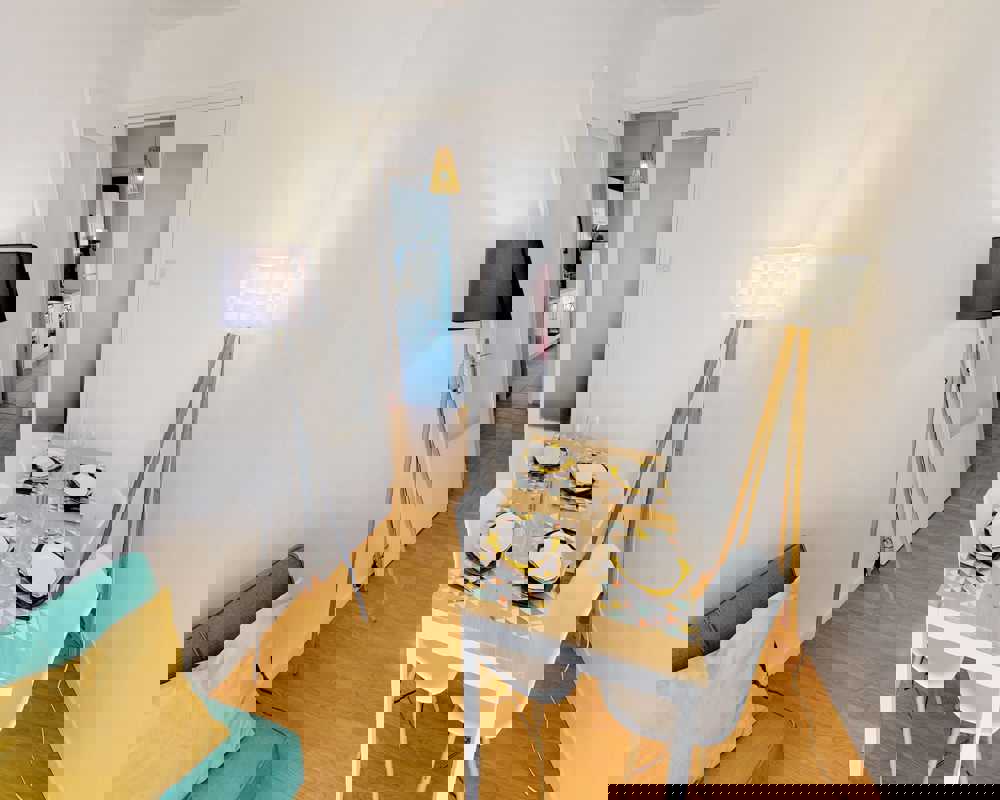 Zona giorno/pranzo luminosa e accogliente con un tavolo in legno per quattro persone, lampade da terra di design e un piccolo divano con cuscini colorati — perfetta per un appartamento compatto.