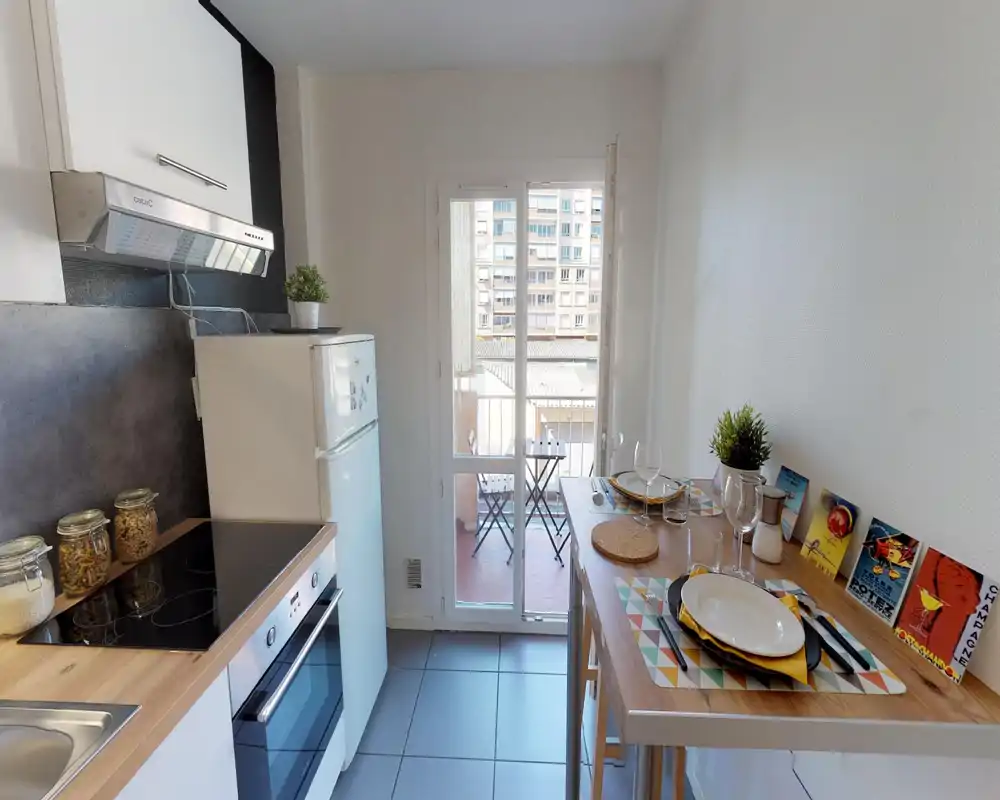 Cucina compatta e luminosa con piano a induzione moderno, forno integrato e piccolo bancone da pranzo per due; porta finestra che si apre su un balcone soleggiato.