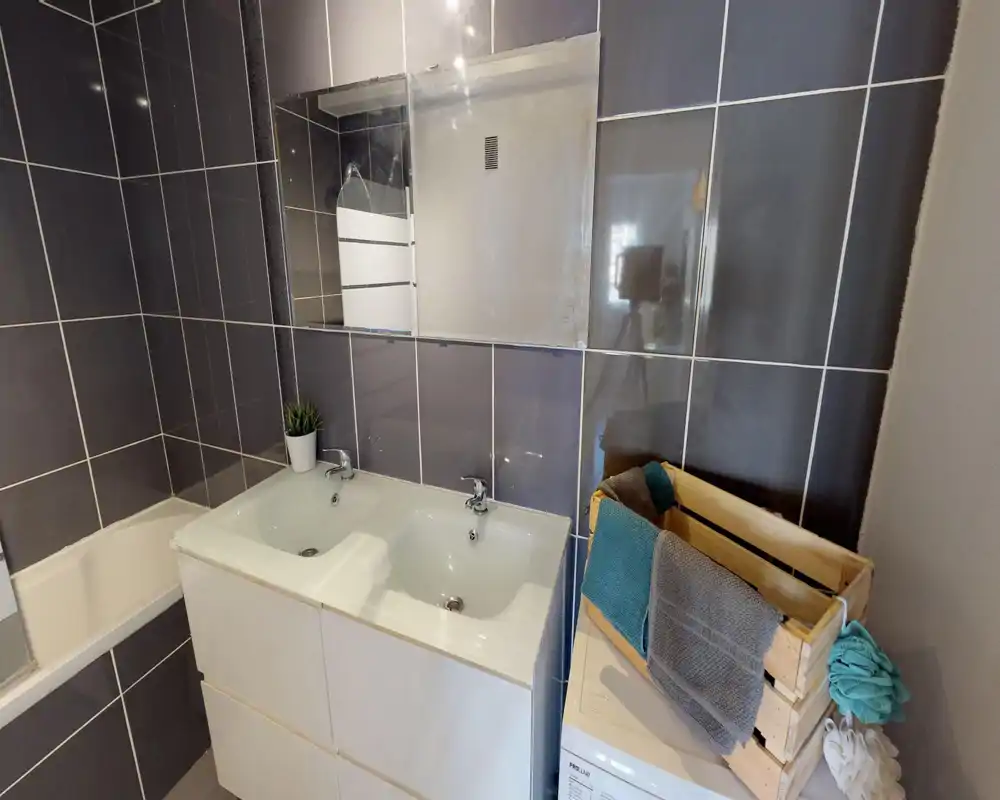 Bagno moderno con lavabo doppio, specchio a parete e piastrelle scure. Lo spazio è ordinato e ben illuminato, con asciugamani piegati e una piccola pianta che aggiungono un tocco curato.