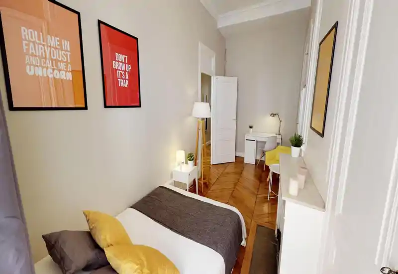 Bright, stylish bedroom with a neatly made double bed, warm parquet flooring, and a small workspace at the far end — cozy decorative pillows and lamps create a welcoming atmosphere.