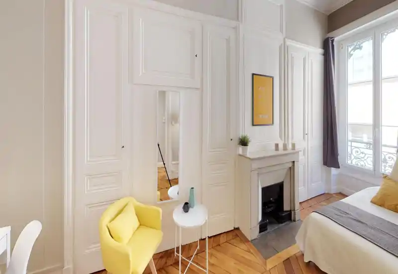 Bright, stylish bedroom corner with classic mouldings and parquet flooring. The image shows a partial bed with warm textiles, a decorative fireplace, a yellow armchair and large window providing abundant natural light.