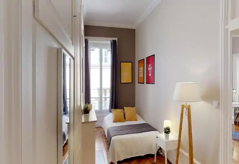 Compact, well-lit bedroom with a neatly made double bed, warm wooden parquet and decorative framed prints. Natural light from the tall window plus bedside lamp creates a welcoming atmosphere ideal for short-term stays.