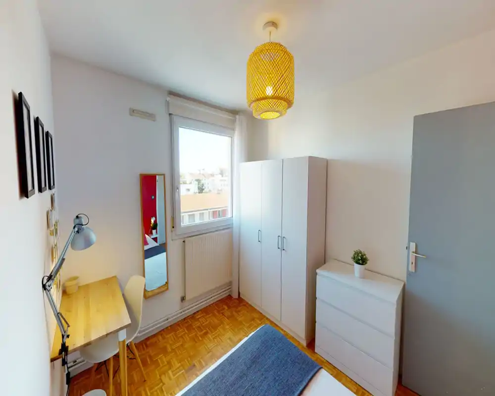 Bright, neatly staged bedroom with a visible double bed, a study desk by the wall and ample storage (wardrobe and chest of drawers). Large window provides natural light and the warm woven ceiling lamp gives a cozy atmosphere.